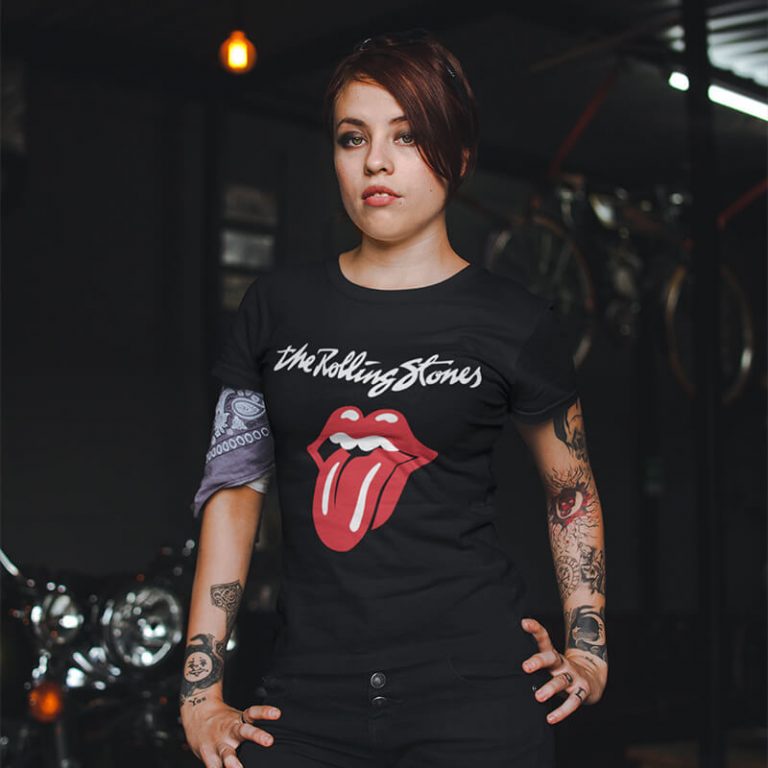 A young woman with tattoos and a dark outfit poses confidently in a dimly lit garage setting. She is wearing a black T-shirt featuring the iconic Rolling Stones logo with a large red tongue and lips graphic. Motorcycles are visible in the background.